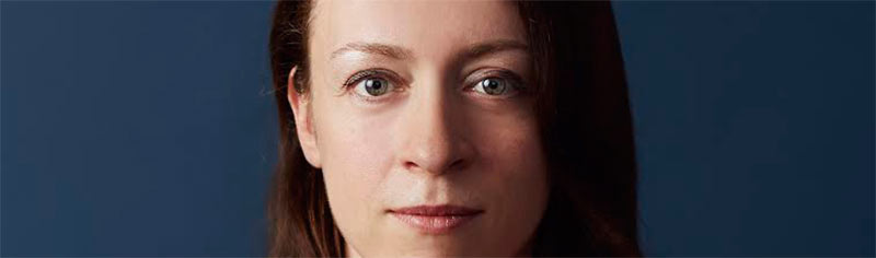 Irina Blok, criadora do mascote atual do Android. Imagem do rosto de uma mulher branca, de cabelos lisos castanhos e olhos verdes de cerca de quarenta anos olhando para a câmera com uma expressão facial neutra. O fundo da imagem é de cor azul escuro sólida.